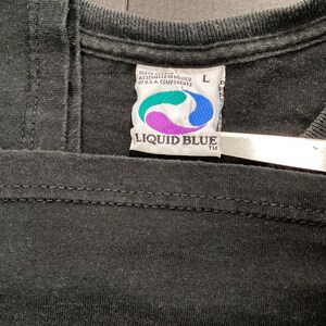 May include: Close-up of a black t-shirt collar with a Liquid Blue label. The label features a teal, purple, and blue design with the brand name. The shirt is size L and made of 100% cotton.