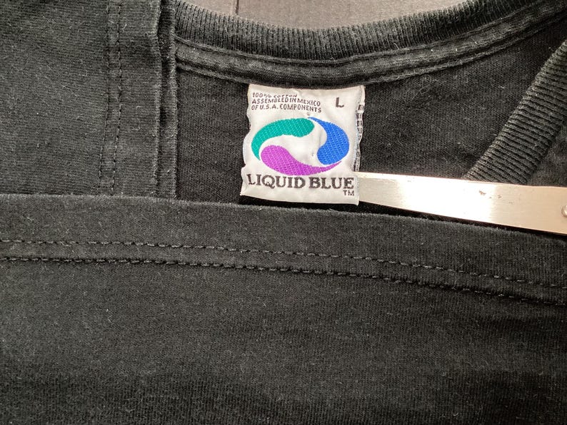 May include: Close-up of a black t-shirt collar with a Liquid Blue label. The label features a teal, purple, and blue design with the brand name. The shirt is size L and made of 100% cotton.
