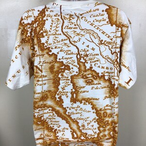 May include: A white t-shirt with a brown vintage map print of Southeast Asia. The map includes the countries of Siam, Cambodia, Cochin China, and more. The map is printed in a faded brown color, giving it a vintage look.