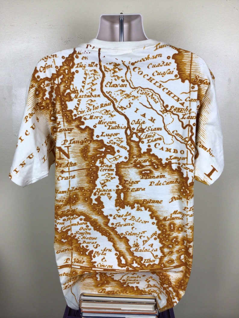 May include: A white t-shirt with a brown vintage map print of Southeast Asia. The map includes the countries of Siam, Cambodia, Cochin China, and more. The map is printed in a faded brown color, giving it a vintage look.