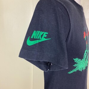 Pu&ograve; includere: T-shirt nero con logo Nike verde sulla manica. La maglietta presenta un design grafico con elementi rossi e verdi. La manica presenta un piccolo strappo vicino alla cucitura. La maglietta &egrave; esposta su un manichino.