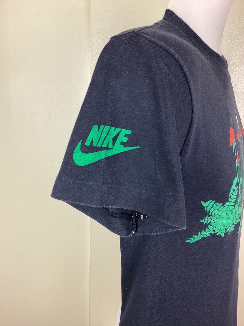 Pu&ograve; includere: T-shirt nero con logo Nike verde sulla manica. La maglietta presenta un design grafico con elementi rossi e verdi. La manica presenta un piccolo strappo vicino alla cucitura. La maglietta &egrave; esposta su un manichino.