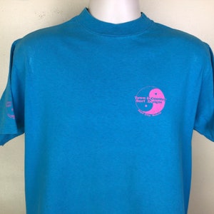 K&ouml;nnte beinhalten: Ein hellblaues T-Shirt mit einem rosafarbenen Yin-Yang-Symbol und dem Text "Town & Country Surf Designs" und "Port City, Hawaii".