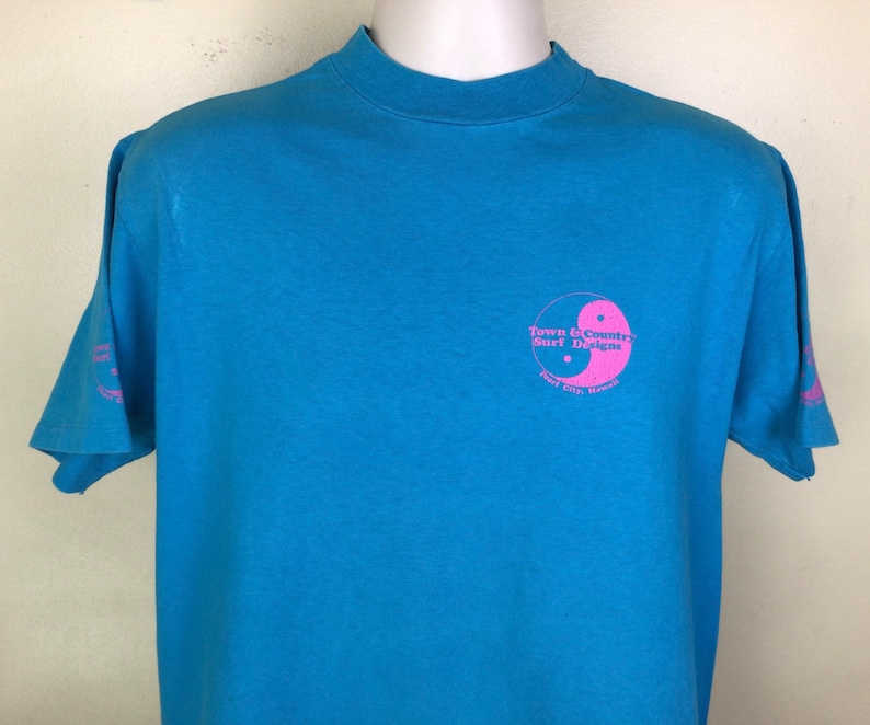 K&ouml;nnte beinhalten: Ein hellblaues T-Shirt mit einem rosafarbenen Yin-Yang-Symbol und dem Text "Town & Country Surf Designs" und "Port City, Hawaii".
