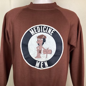 Puede incluir: Sudadera marrón con cuello redondo y mangas largas. La parte delantera presenta un gráfico circular con las palabras "MEDICINE MEN" y una figura de dibujos animados nativa americana sosteniendo un letrero que dice "Class of '82".