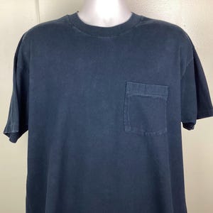 May include: A navy blue, short-sleeved t-shirt with a chest pocket. The shirt has a crew neck and a slightly faded appearance. The fabric appears to be a soft cotton blend, suitable for casual wear.