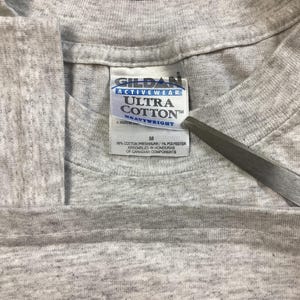 May include: A gray Gildan Ultra Cotton Heavyweight t-shirt tag with the text "Gildan Activewear Ultra Cotton Heavyweight" and "M" for size. The tag is made of white fabric with black text.