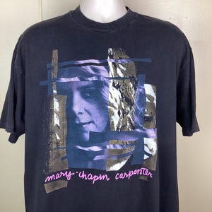 May include: Navy blue t-shirt with a graphic of a woman's face in blue and grey tones. The text "mary-chapin carpenter" is printed in pink cursive below the image. The shirt has short sleeves and a crew neck.