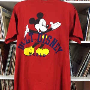 K&ouml;nnte beinhalten: Rotes T-Shirt mit einem Cartoon-Bild von Mickey Mouse, der gelbe Schuhe tr&auml;gt, und dem Text "Walt Disney World" in Blau.