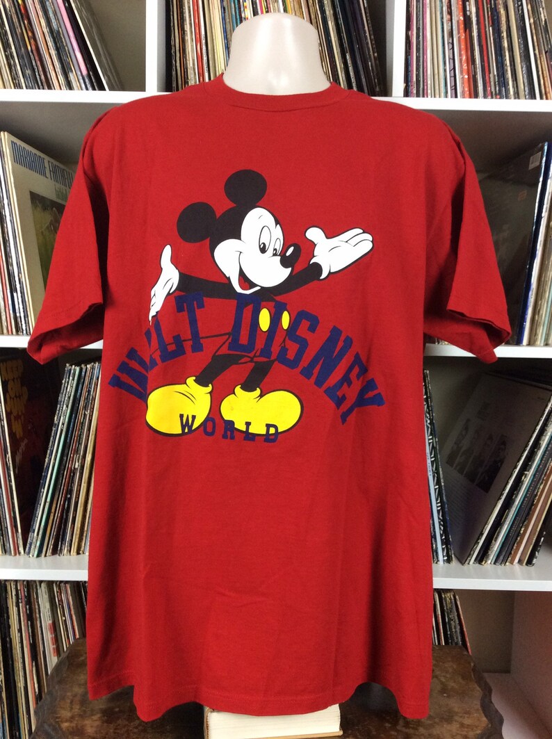 K&ouml;nnte beinhalten: Rotes T-Shirt mit einem Cartoon-Bild von Mickey Mouse, der gelbe Schuhe tr&auml;gt, und dem Text "Walt Disney World" in Blau.