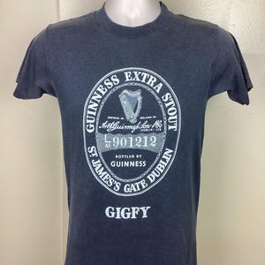 May include: A dark blue t-shirt with a white print of the Guinness Extra Stout logo. The logo includes the text "Guinness Extra Stout", "Brewed in Ireland by Arthur Guinness Son & Co. (Dublin) Ltd", "L 901212", "Bottled by Guinness", "St. James's Gate Dublin", and "Gigfy".