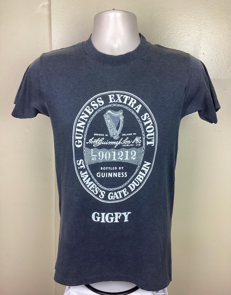 May include: A dark blue t-shirt with a white print of the Guinness Extra Stout logo. The logo includes the text "Guinness Extra Stout", "Brewed in Ireland by Arthur Guinness Son & Co. (Dublin) Ltd", "L 901212", "Bottled by Guinness", "St. James's Gate Dublin", and "Gigfy".