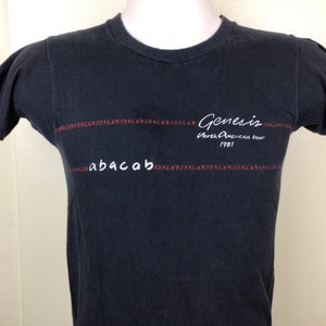 May include: Black t-shirt with white text that reads "Genesis North American Tour 1981" and "abacab" in a red and white pattern.