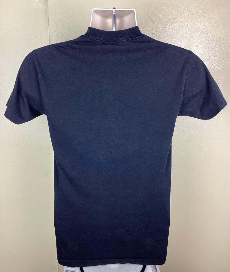 Pu&ograve; includere: T-shirt nero tinta unita a maniche corte, presentato su un manichino. La maglietta ha un girocollo e un design semplice e classico. Il tessuto sembra essere un morbido misto cotone, adatto per abbigliamento casual.