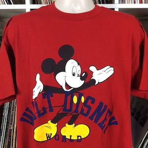 K&ouml;nnte beinhalten: Rotes T-Shirt mit einem Cartoon-Bild von Mickey Mouse vor einem blauen und gelben "Walt Disney World"-Logo.