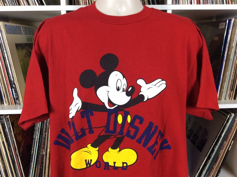 K&ouml;nnte beinhalten: Rotes T-Shirt mit einem Cartoon-Bild von Mickey Mouse vor einem blauen und gelben "Walt Disney World"-Logo.