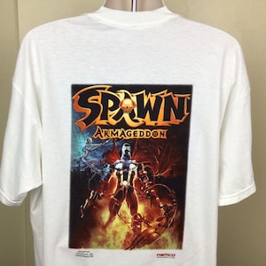 May include: White t-shirt with a large graphic print of Spawn: Armageddon. The graphic features Spawn in a red cape and black suit, with a skull on his chest. The text "Spawn: Armageddon" is in gold letters.