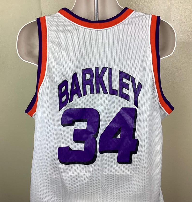 May include: White basketball jersey with the name "BARKLEY" and the number "34" in purple. The jersey has orange and purple trim around the neck and arm openings. The jersey is displayed on a mannequin.