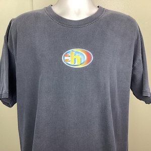 Pode incluir: Uma camiseta cinza escuro com gola redonda e mangas curtas. A camiseta apresenta um logotipo colorido em forma oval na frente. O logotipo tem um "H" amarelo e laranja dentro de um oval azul e amarelo.