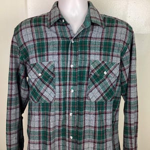 Pode incluir: Uma camisa de manga comprida xadrez em verde, cinza e vermelho. A camisa tem colarinho de botões e dois bolsos no peito.