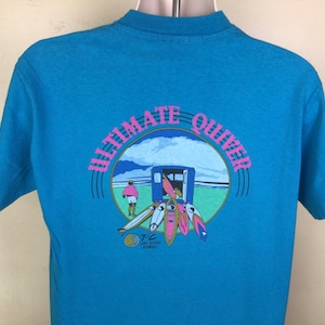 K&ouml;nnte beinhalten: Ein blaues T-Shirt mit einem Aufdruck eines Surfshops mit dem Text "Ultimate Quiver" in einem Kreis. Der Shop befindet sich an einem Strand mit einer Person, die im Hintergrund geht. Der Text "TC Surf Designs Hawaii" befindet sich unter dem Kreis.