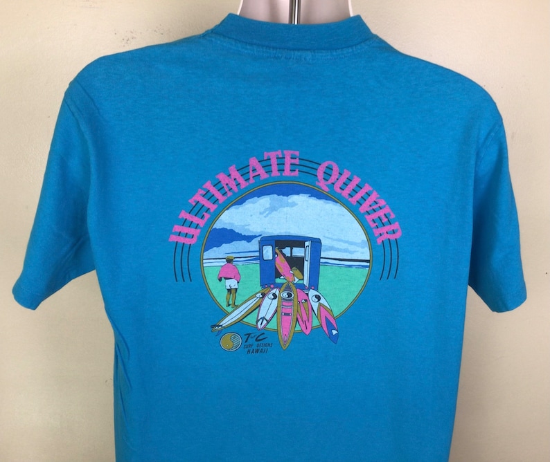 K&ouml;nnte beinhalten: Ein blaues T-Shirt mit einem Aufdruck eines Surfshops mit dem Text "Ultimate Quiver" in einem Kreis. Der Shop befindet sich an einem Strand mit einer Person, die im Hintergrund geht. Der Text "TC Surf Designs Hawaii" befindet sich unter dem Kreis.