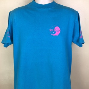 K&ouml;nnte beinhalten: Ein t&uuml;rkisfarbenes T-Shirt mit einem rosafarbenen Yin-Yang-Symbol und dem Text "Town & Country Surf Designs" und "Kailua City, Hawaii".
