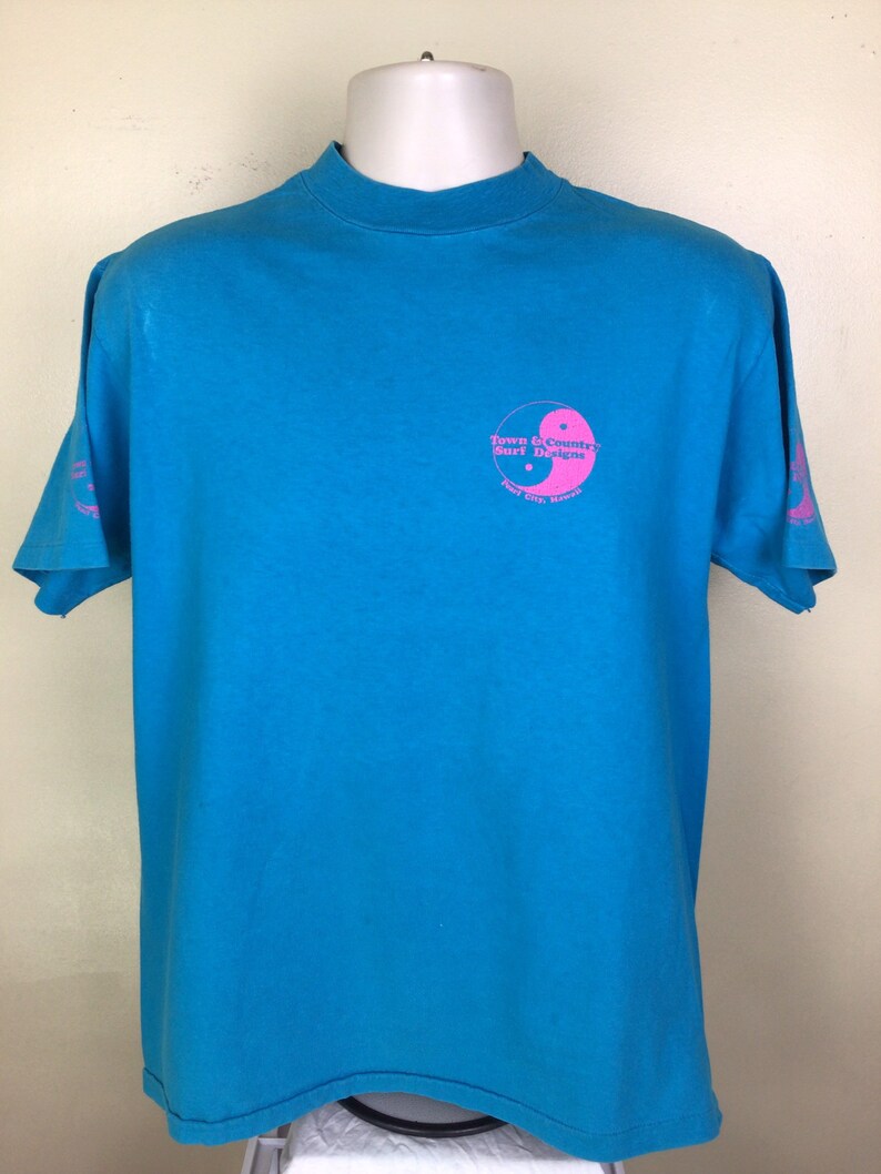 K&ouml;nnte beinhalten: Ein t&uuml;rkisfarbenes T-Shirt mit einem rosafarbenen Yin-Yang-Symbol und dem Text "Town & Country Surf Designs" und "Kailua City, Hawaii".