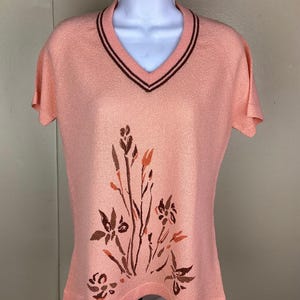May include: A peach-colored short-sleeved sweater with a V-neck and a floral design. The neckline is trimmed with two dark brown stripes. The floral design is in shades of brown and orange.