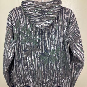 May include: A long-sleeve hooded sweatshirt featuring a Realtree camouflage pattern with brown, gray, and white bark textures and green leaf accents. The garment has a solid black ribbed hem and cuffs, a drawstring hood, and a full-length front zipper.
