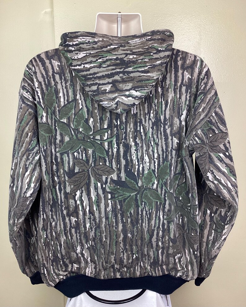May include: A long-sleeve hooded sweatshirt featuring a Realtree camouflage pattern with brown, gray, and white bark textures and green leaf accents. The garment has a solid black ribbed hem and cuffs, a drawstring hood, and a full-length front zipper.