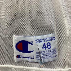 May include: Close-up of a white mesh jersey with a Champion tag. The tag features the Champion logo in blue and red, the number 48, and the text "IT TAKES A LITTLE MORE TO MAKE A CHAMPION".