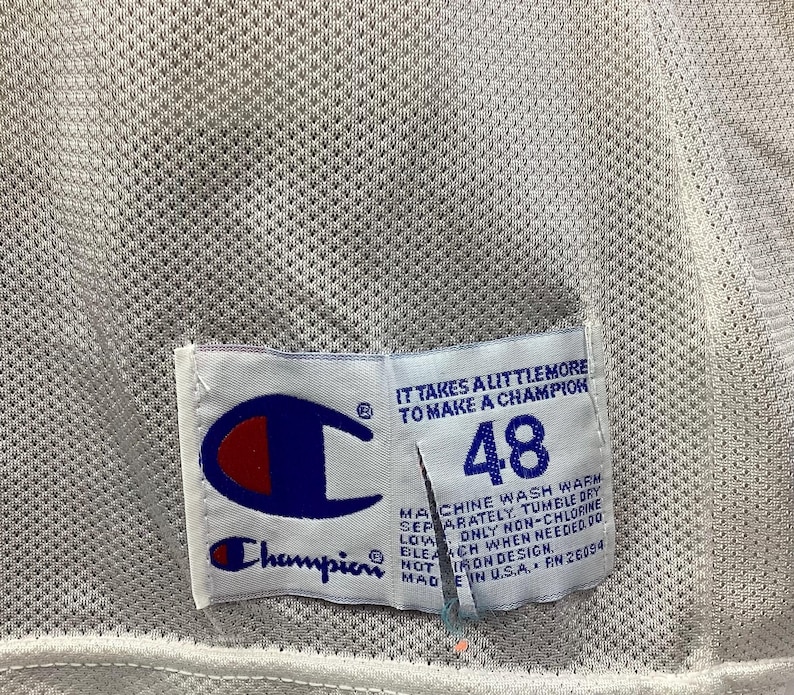 May include: Close-up of a white mesh jersey with a Champion tag. The tag features the Champion logo in blue and red, the number 48, and the text "IT TAKES A LITTLE MORE TO MAKE A CHAMPION".