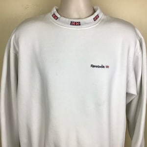 Op de afbeelding: Witte Reebok-trui met een mock neck en een klein geborduurd Union Jack-vlaggetje op de kraag. De trui heeft het woord "Reebok" in het zwart geborduurd op de voorkant.