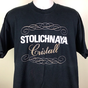 以下が含まれることがあります： 黒いTシャツに、金色のフォントで「Stolichnaya Cristal」という白いロゴが、装飾的な渦巻き模様で描かれている。