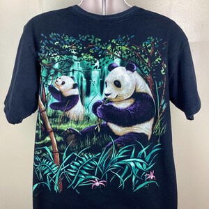 May include: Black t-shirt with a colourful graphic of two pandas in a bamboo forest. The pandas are depicted with black and white fur and purple accents, amidst green foliage and pink flowers.