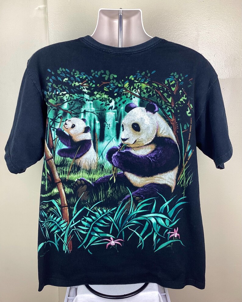 May include: Black t-shirt with a colourful graphic of two pandas in a bamboo forest. The pandas are depicted with black and white fur and purple accents, amidst green foliage and pink flowers.