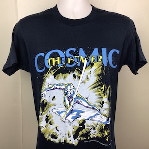 Pu&ograve; includere: Maglietta nera con una grafica del Silver Surfer, un supereroe dei Marvel Comics, che vola nello spazio. Il testo "COSMIC THE POWER" &egrave; stampato in blu sopra la grafica.
