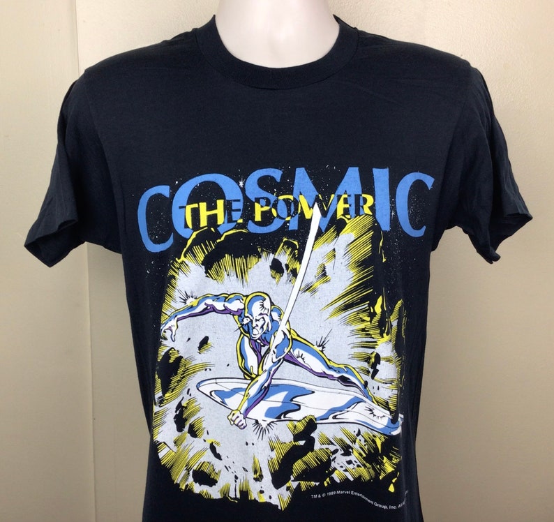 Pu&ograve; includere: Maglietta nera con una grafica del Silver Surfer, un supereroe dei Marvel Comics, che vola nello spazio. Il testo "COSMIC THE POWER" &egrave; stampato in blu sopra la grafica.