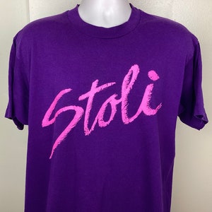 以下が含まれることがあります： 前面にピンクの「Stoli」ロゴがプリントされたパープルのTシャツです。