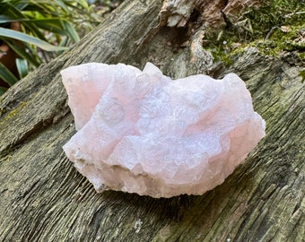 Puede incluir: Un gran cúmulo de cristales rosa con una superficie rugosa y texturizada. El cristal está sentado sobre un trozo de madera desgastada.
