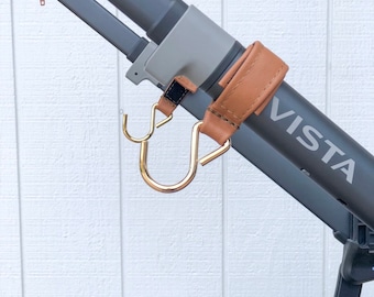 May include: A brown leather strap with a gold metal hook attached to a gray stroller handle. The stroller handle has the word "VISTA" printed on it.