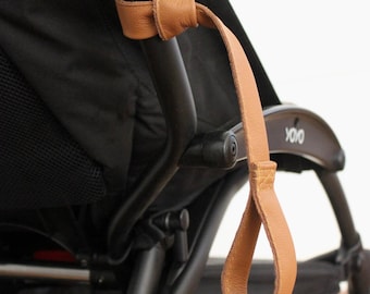 Può includere: Un cinturino in pelle marrone è attaccato alla maniglia di un passeggino nero. Il passeggino ha un sedile in tessuto nero e un telaio in metallo nero. Il passeggino è etichettato "YOYO" sul lato.