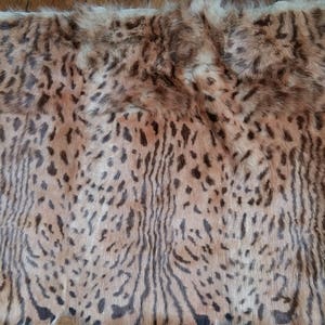 May include: A rectangular piece of animal fur with a spotted pattern in shades of brown and black. The fur has a soft, textured appearance, with a lighter tan base and darker spots. The edges of the fur are uneven.