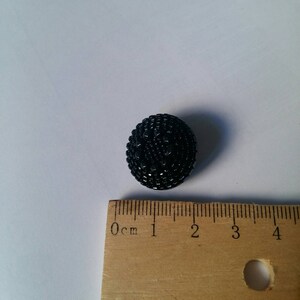 May include: A black, spherical button with a textured surface. The button is approximately 1 inch in diameter. The button is placed next to a wooden ruler.
