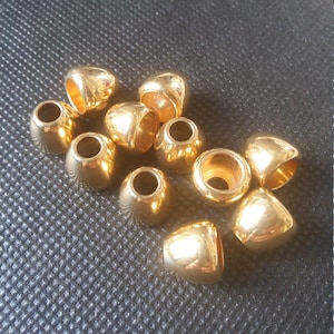 Peut inclure: Un groupe de 10 perles en métal de couleur dorée avec un dessus rond légèrement pointu et un trou au centre. Les perles sont dispersées sur une surface noire texturée.