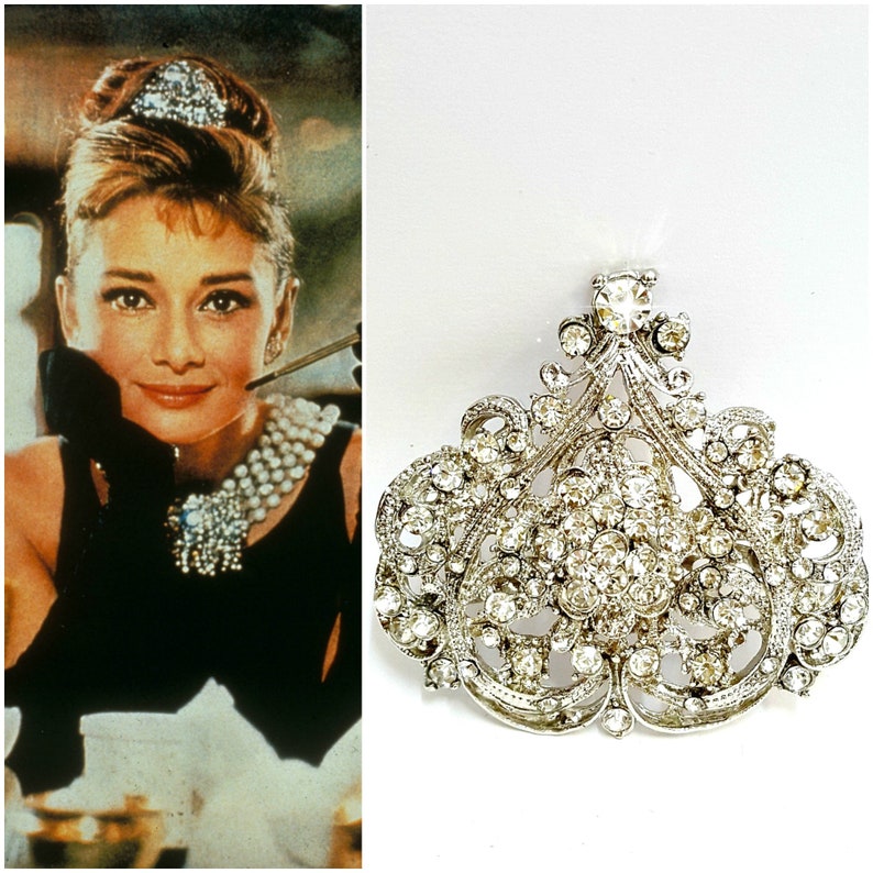 Pu&ograve; includere: Un fermaglio in argento e cristallo con un grande design elaborato. Il fermaglio ha la forma di una lacrima e presenta un gruppo di cristalli trasparenti al centro.
