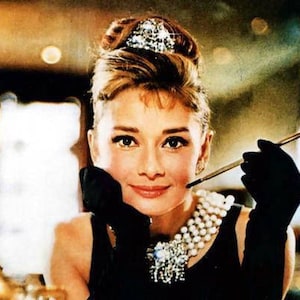 Pu&ograve; includere: Una donna con un'acconciatura classica e una collana di perle tiene un porta sigarette nella mano destra. Indossa un abito nero e guanti neri. Lo sfondo &egrave; sfocato.