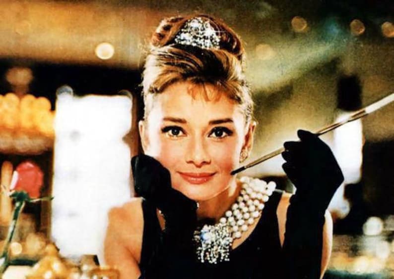 Pu&ograve; includere: Una donna con un'acconciatura classica e una collana di perle tiene un porta sigarette nella mano destra. Indossa un abito nero e guanti neri. Lo sfondo &egrave; sfocato.