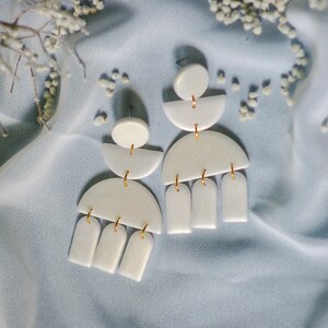 Peut inclure: Une paire de boucles d'oreilles géométriques blanches avec des accents dorés. Les boucles d'oreilles sont en argile polymère et présentent un demi-cercle, un cercle et trois formes rectangulaires.